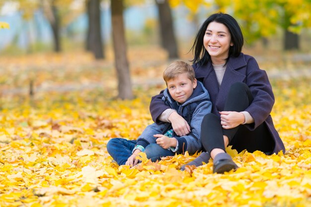 Szczęśliwa, zadowolona mama i syn siedzą w miejskim parku wśród żółtych jesiennych liści.