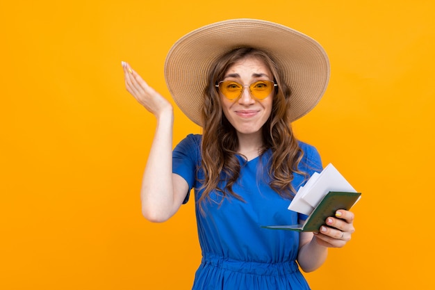 szczęśliwa zaskoczona młoda kobieta w niebieskiej sukience, słomkowym kapeluszu i okularach przeciwsłonecznych z paszportem i biletami na wakacje na żółto
