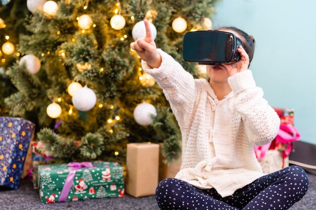 Szczęśliwa zdumiona dziewczynka nosząca okulary wirtualnej rzeczywistości, oglądając filmy lub grając w gry wideo, na białym tle na tle Bożego Narodzenia. Wesołe zdziwione dziecko patrzące w okulary VR i gestykulujące swoim