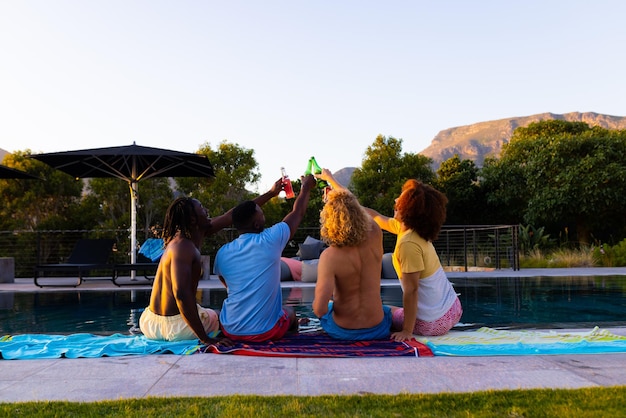 Szczęśliwa, zróżnicowana grupa przyjaciół urządzających imprezę przy basenie, pijących piwo i wznoszących toast w ogrodzie. Styl życia, przyjaźń i impreza, lato, słońce, bez zmian.
