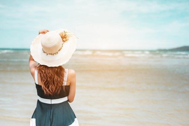 Szczęśliwe azjatyckie kobiety z trzymającym kapelusz stoją, aby otrzymać wiatr i cieszyć się życiem na wakacjach na plaży. plaża, lato, styl windy, pozytywny nastrój, podróże, koncepcja relaksu.