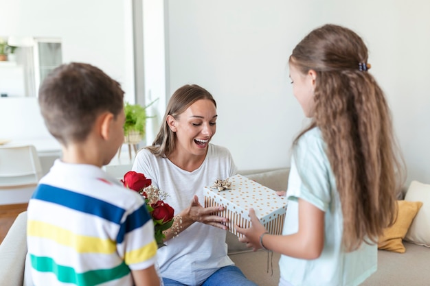 Szczęśliwe Dzieci Dające W Prezencie Kwiaty Matce. Szczęśliwego Dnia Matki! Dzieci Chłopiec I Dziewczynka Gratulują Uśmiechniętej Mamie, Wręczają Jej Bukiet Kwiatów Z Róż I Pudełko Prezentowe Podczas świątecznych Uroczystości