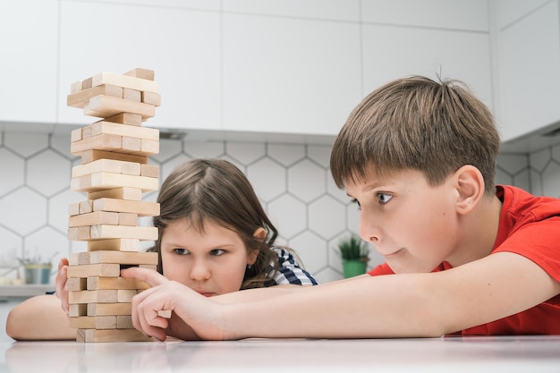 Szczęśliwe dzieci szkolne bawią się w wieżę siedząc przy stole w kuchni Skoncentrowany chłopiec i dziewczynka budują wieżę z małych drewnianych klocków