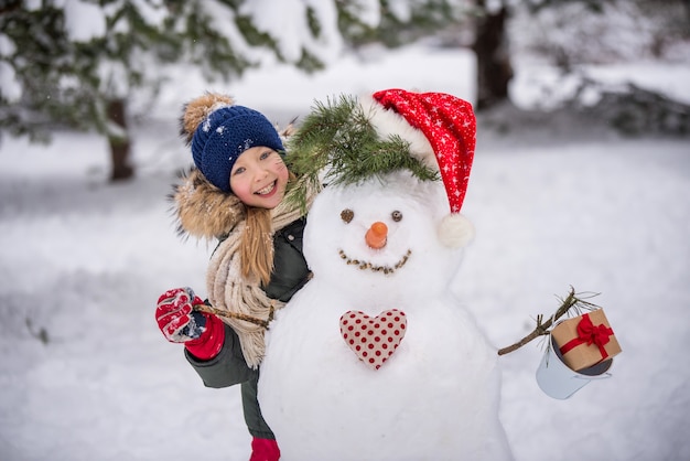 Szczęśliwe dziecko blond śliczna dziewczyna gra z bałwanem na śnieżnym zimowym spacerze