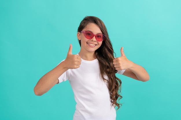 Szczęśliwe dziecko długie kręcone włosy w okularach przeciwsłonecznych w stylu casual na niebieskim tle kciuk w górę w letnim stylu
