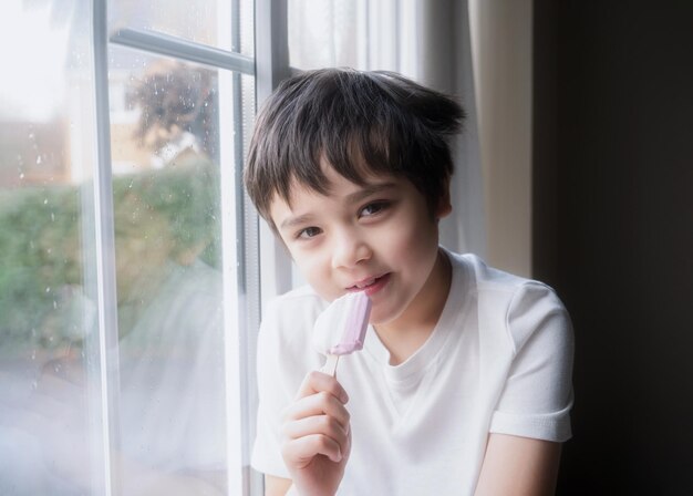 Szczęśliwe dziecko jedzące lody Portret przystojnego chłopca siedzącego przy oknie i jejącego przekąskę Dziecko z uśmiechniętą twarzą relaksujące się w domu Dziecko patrzące na kamerę