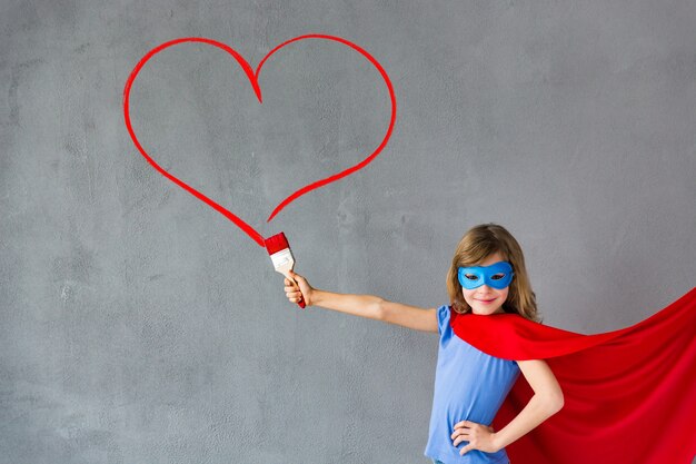 Zdjęcie szczęśliwe dziecko malarstwo duże czerwone serce na ścianie. zabawna dziewczyna superbohatera gra w domu. karta walentynki. koncepcja remontowo-projektowa