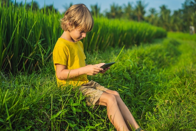 Szczęśliwe dziecko siedzi na polu trzymając tablet Chłopiec siedzi na trawie w słoneczny dzień Domowa nauka lub gra na tablecie