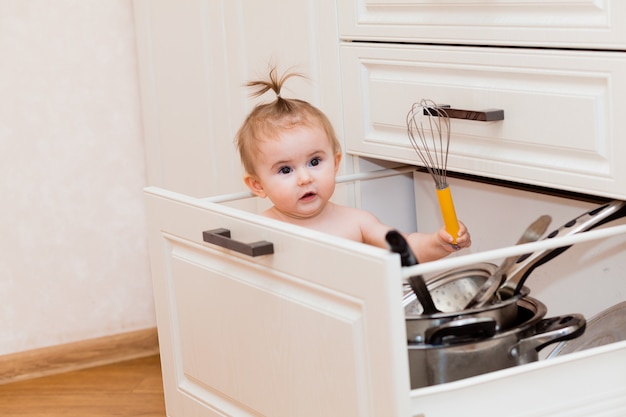 Szczęśliwe dziecko siedzi w szufladzie kuchennej z garnkami i śmiejąc się. Portret malucha w białej kuchni.
