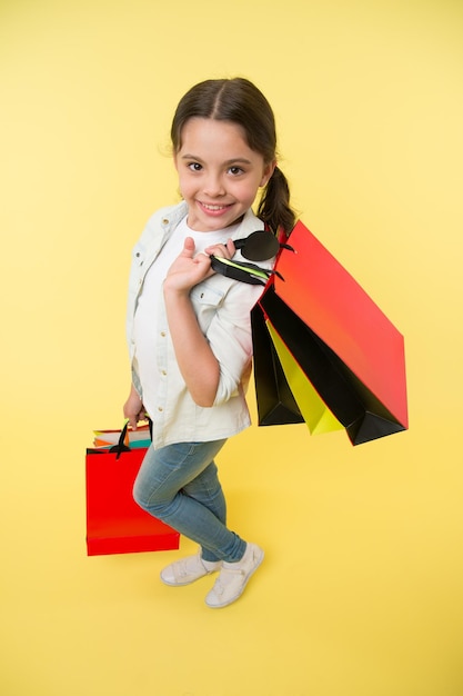 Szczęśliwe dziecko trzyma torby na zakupy na żółtym tle Mały zakupoholiczka uśmiech z papierowymi torbami Dziewczyna shopper przygotowuje się do mody urodzinowej Koncepcja czarnego piątku Zabawa na zakupach Duża wyprzedaż