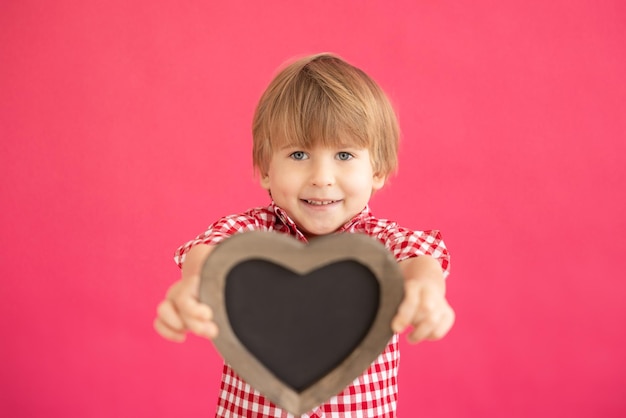 Szczęśliwe dziecko trzymając tablica w kształcie serca puste. Portret uśmiechniętego dzieciaka na różowym tle. Koncepcja wakacje walentynki