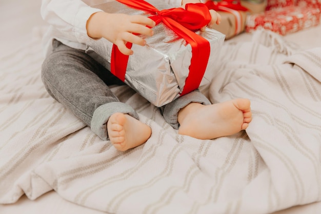 Szczęśliwe dziecko w czapce Mikołaja siedzi na podłodze w pobliżu choinki i trzyma w rękach pudełko z prezentem. gołe obcasy z bliska