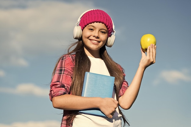 Szczęśliwe dziecko w swobodnym stylu trzyma jabłko i książkę szkolną słuchając dźwięku w słuchawkach wiedzy