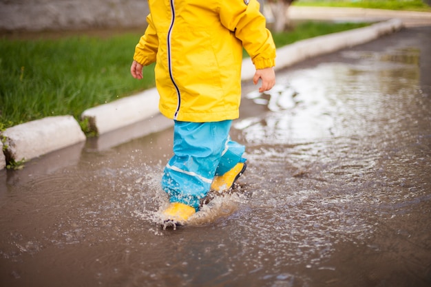 szczęśliwe dziecko w żółtych butach, żółtej kurtce i niebieskich jasnych spodniach przechodzi przez kałuże i rozpyla we wszystkich kierunkach. Wiosenna deszczowa pogoda i szczęśliwe dzieciństwo.