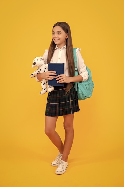 Szczęśliwe dziecko z zabawką w plecaku i książką w mundurku szkolnym pełnej długości na żółtym tle studium