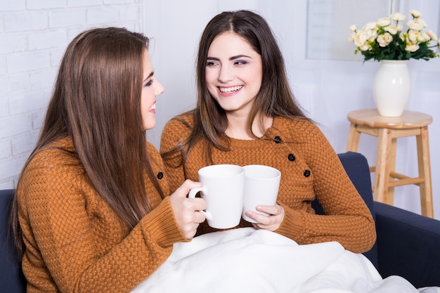 Szczęśliwe dziewczyny rozmawiają i piją kawę lub herbatę siedząc na kanapie w domu