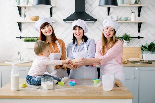 Szczęśliwe kobiety w białych fartuchach piec wpólnie i trzyma ciasto w rękach w lekkiej kuchni. Mała dziewczynka pomaga robić babeczki razem z matką, ciotką i babcią