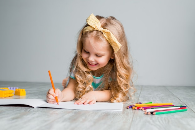 Szczęśliwe małe dziecko, urocze blond dziecko leży wygodnie na drewnianej podłodze, rysując na papierze z kolorowymi ołówkami