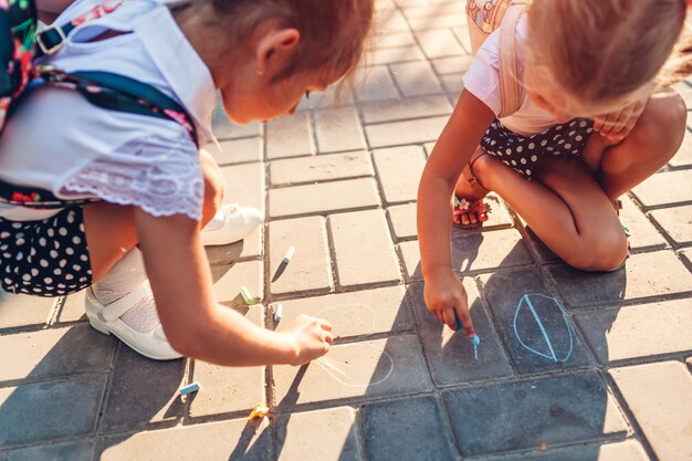 Szczęśliwe Małe Dziewczynki Jest Ubranym Plecaki I Rysuje Kredą Outdoors Szkołą Podstawową.