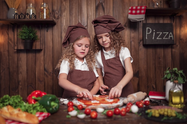 Szczęśliwe małe dziewczynki z wałkiem i szpachelką uśmiechnięte i patrząc na kamery, przygotowując wspólnie pyszną pizzę w przytulnej kuchni