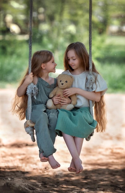 Szczęśliwe roześmiane dzieci dziewczyny siostry z długimi włosami cieszące się przejażdżką huśtawką z zabawką pluszowego misia w słoneczny letni dzień