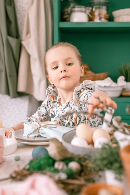 Szczęśliwe, szczere małe czteroletnie dziecko bawi się gotowymi wiosennymi świętami wielkanocnymi w domowej kuchni pomaga matce dekorować świąteczny stół z jajkami wielkanocnymi i piekarnią oraz słodyczami na lunch lub kolację