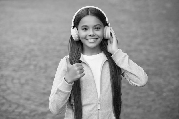 Szczęśliwe uśmiechnięte dziecko lubi słuchać muzyki w słuchawkach dziecko słuchać piosenki spacery na świeżym powietrzu z ulubioną melodią edukacja online koncepcja dzieciństwo szczęście e-learning studentka dziewczyna kciuk w górę