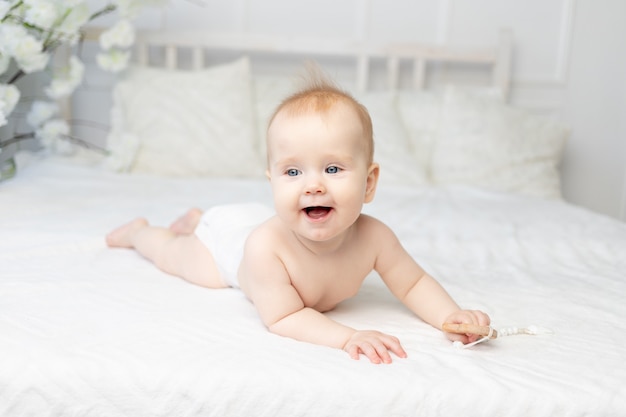 Szczęśliwe uśmiechnięte dziecko w pieluchach na białym bawełnianym łóżku w domu w wieku sześciu miesięcy
