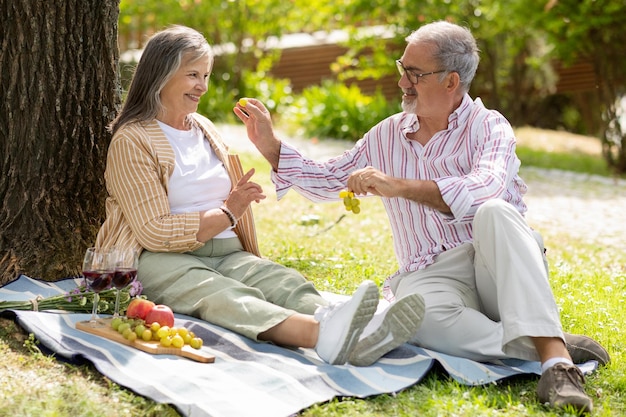 Szczęśliwego starszego Europejczyka karmiącego winogrona pani, ciesz się romantycznym obiadem na randce w parku, baw się dobrze