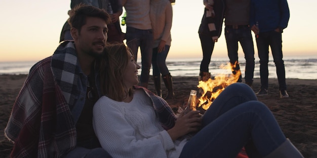 Szczęśliwi beztroscy młodzi przyjaciele bawią się i piją piwo przy ognisku na plaży, gdy słońce zaczyna zachodzić