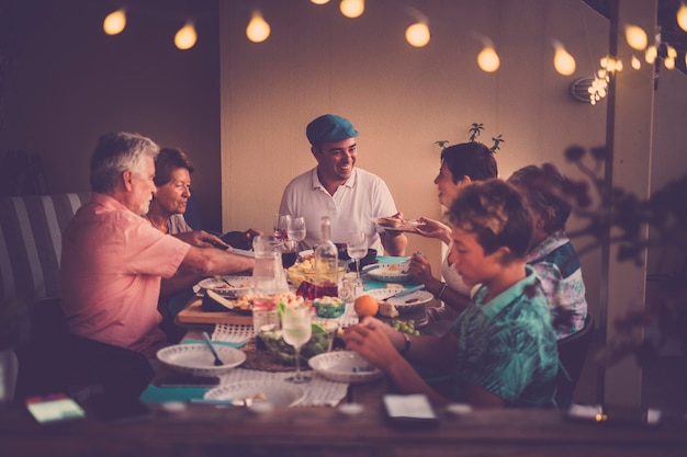 Szczęśliwi I Przyjaźni Ludzie Jedzący Wspólnie Kolację W Domu Lub Restauracji W Przyjaźni - Rodziny W Różnym Wieku I W Różnym Wieku, Mężczyźni I Kobiety, Cieszący Się Jedzeniem Przy Stole