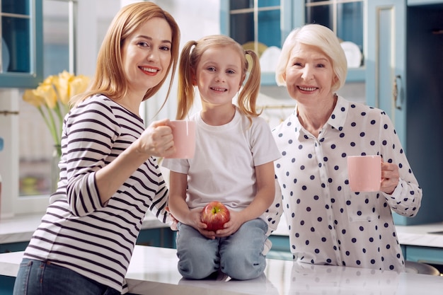 Szczęśliwi Krewni. Urocza Mała Dziewczynka Siedzi Na Kuchennym Blacie I Trzyma Jabłko, Podczas Gdy Jej Optymistyczna Mama I Babcia Stoją Obok Niej I Trzymają Filiżanki Z Kawą