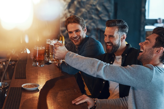 Szczęśliwi młodzi mężczyźni w luźnych ubraniach, wznoszący toast z piwem i śmiejący się, siedząc w pubie