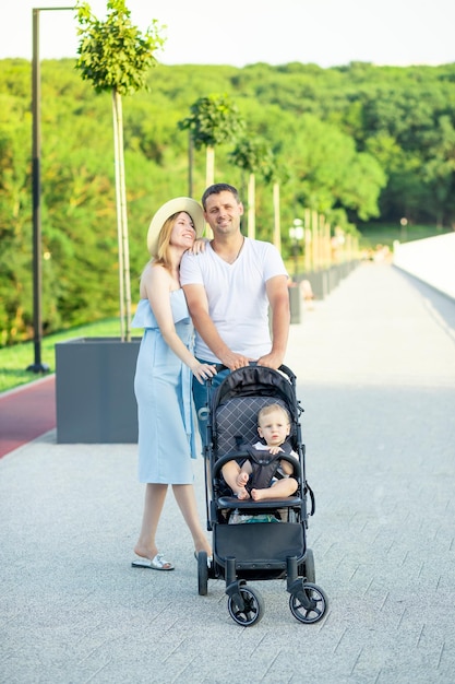 Szczęśliwi młodzi rodzice mama i tata spacerują z dzieckiem w wózku w parku w kapeluszu latem w zachodzącym słońcu i uśmiechają się, bawiąc się