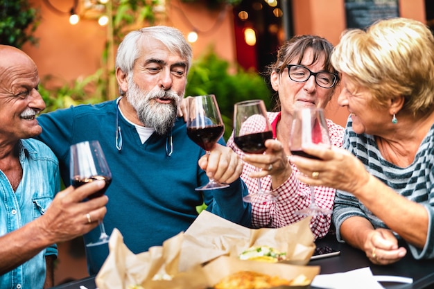 Szczęśliwi seniorzy bawią się opiekaniem czerwonego wina podczas kolacji w ogrodzie