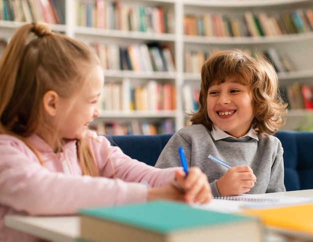 Szczęśliwi uczniowie, przyjaciele siedzący przy stole w klasie w szkole podstawowej, rozmawiający i uśmiechający się