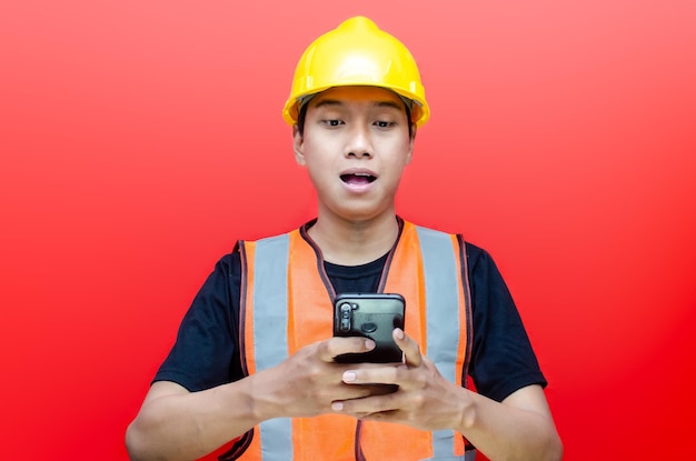 szczęśliwie zszokowany azjatycki mężczyzna robotnik budowlany lub inżynier w kasku ochronnym i kamizelce trzymającej telefon