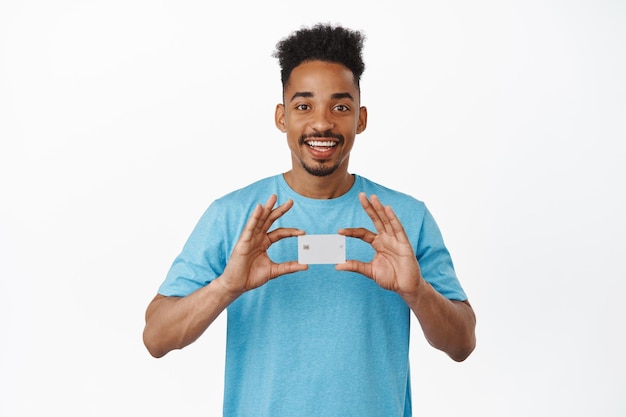 Szczęśliwy afroamerykanin pokazujący kartę kredytową, uśmiechający się zadowolony, polecający bank, robiący zakupy w sklepie, stojący na białym tle