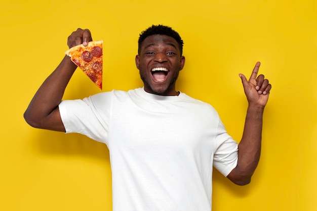 szczęśliwy Afroamerykanin w białej koszulce trzyma kawałek pizzy i pokazuje rękę na bok