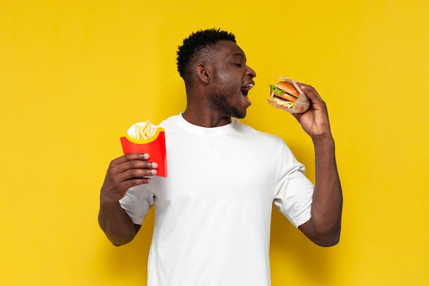Szczęśliwy Afroamerykanin w białej koszulce trzymający duży hamburger i frytki i uśmiechający się