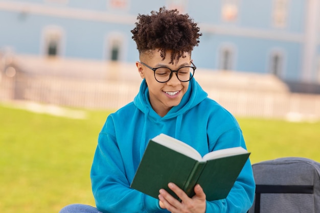 Szczęśliwy afroamerykański student w okularach czytający książkę na zewnątrz