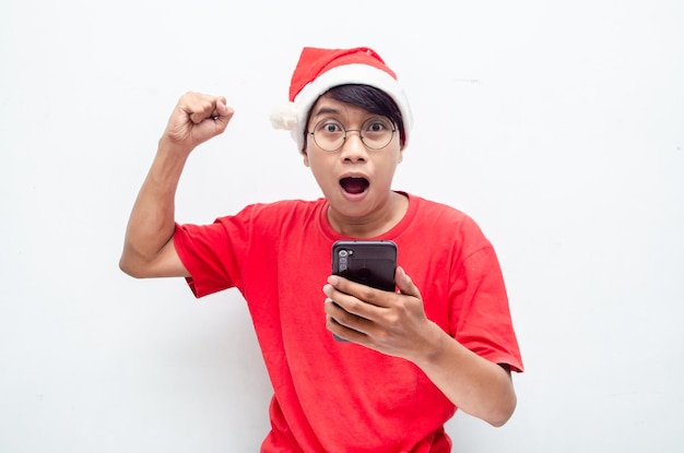 szczęśliwy atrakcyjny azjatycki człowiek w czerwonych świątecznych ubraniach tematycznych zszokowany szczęśliwie trzymając telefon.