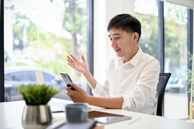 Szczęśliwy azjatycki biznesmen używający smartfona siedząc przy biurku