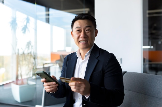 Szczęśliwy azjatycki mężczyzna robi zakupy online biznesmen w biurze zakupy w sklepie internetowym za pomocą karty kredytowej zadowolony z pożyczki