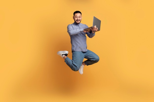 Szczęśliwy azjatycki mężczyzna z laptopem skacze nad żółtym tłem
