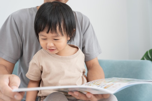 Szczęśliwy azjatycki ojciec odpoczywa i czyta książkę z dzieckiem czas razem w domu rodzic siedzi na kanapie z córką i czyta historię uczyć się rozwoju opieka nad dzieckiem śmiech edukacja praktyka opowiadania historii