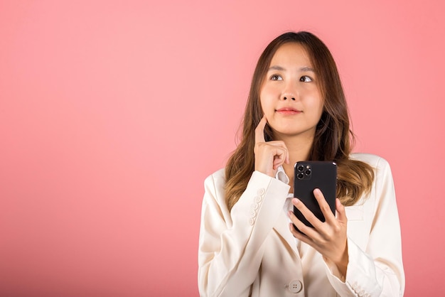 Szczęśliwy Azjatycki Portret Piękna śliczna Młoda Kobieta Podekscytowana Uśmiechnięta Trzymając Telefon Komórkowy I Myśląc, że Pomysł Palcem Dotyka Twarzy, Studio Strzał Odizolowane Na Różowym Tle, Kobieta Za Pomocą Smartfona Wykonującego Gest