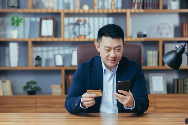 Szczęśliwy azjatycki zakupy online siedzący w biurze mężczyzna korzystający z aplikacji telefonu i karty bankowej biznesmen korzystający z zakupów online