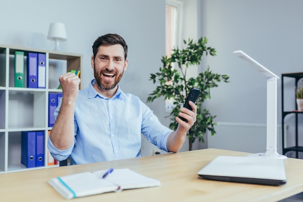 Szczęśliwy biznesmen czytający dobre wieści z telefonu człowieka pracującego w biurze