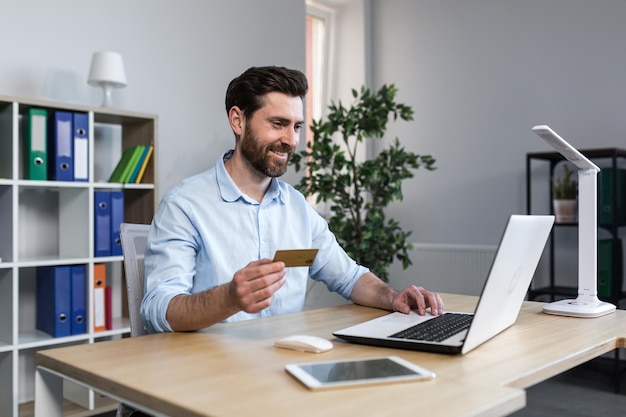 Szczęśliwy biznesmen robi zakupy online pracując w biurze mąż trzyma kartę kredytową i korzysta z laptopa